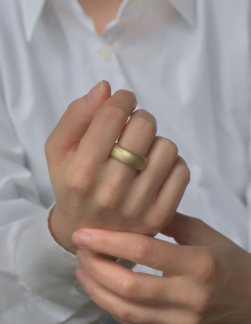 Two-piece gold ring with a matt surface and an oval cross-sectional profile, worn on the middle finger of the left hand.