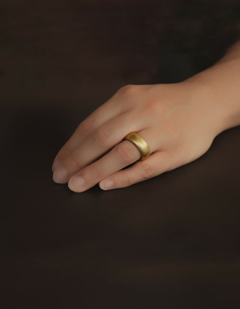 Two-piece gold ring with a matt surface and an oval cross-sectional profile, worn on the ring finger of the left hand.