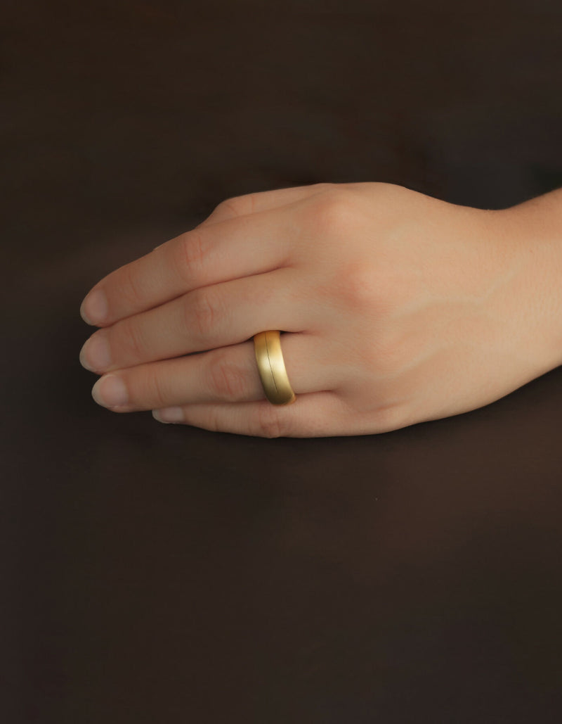 Two-piece gold ring with a matt surface and an oval cross-sectional profile, worn on the ring finger of the left hand.