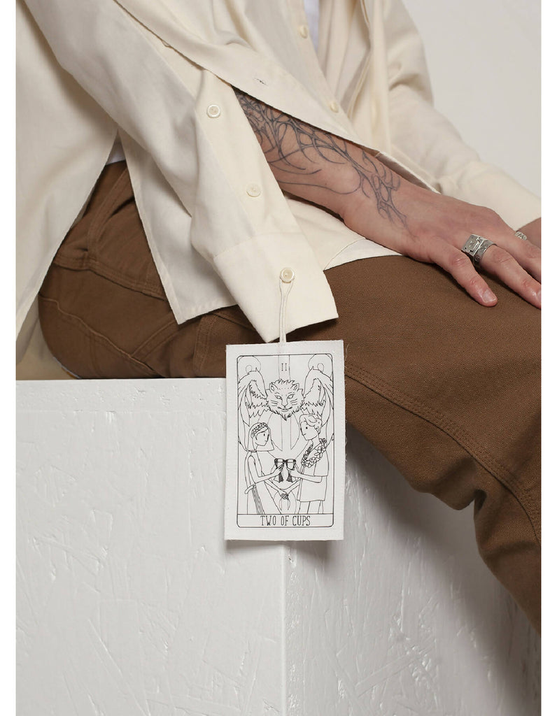 Clément is wearing the White Canvas ESSENTIAL SHIRT styled with THE TWO OF CUPS tag made from leftover deadstock white cotton poplin. The black and white tag has an exclusive design inspired by tarot cards drawn by the artist Maria Robla and is printed in collaboration with RoStudio. Wear the tag alone or attach it to the essential pieces to personalize them. The tag is sold with key holder in recycled plastic.